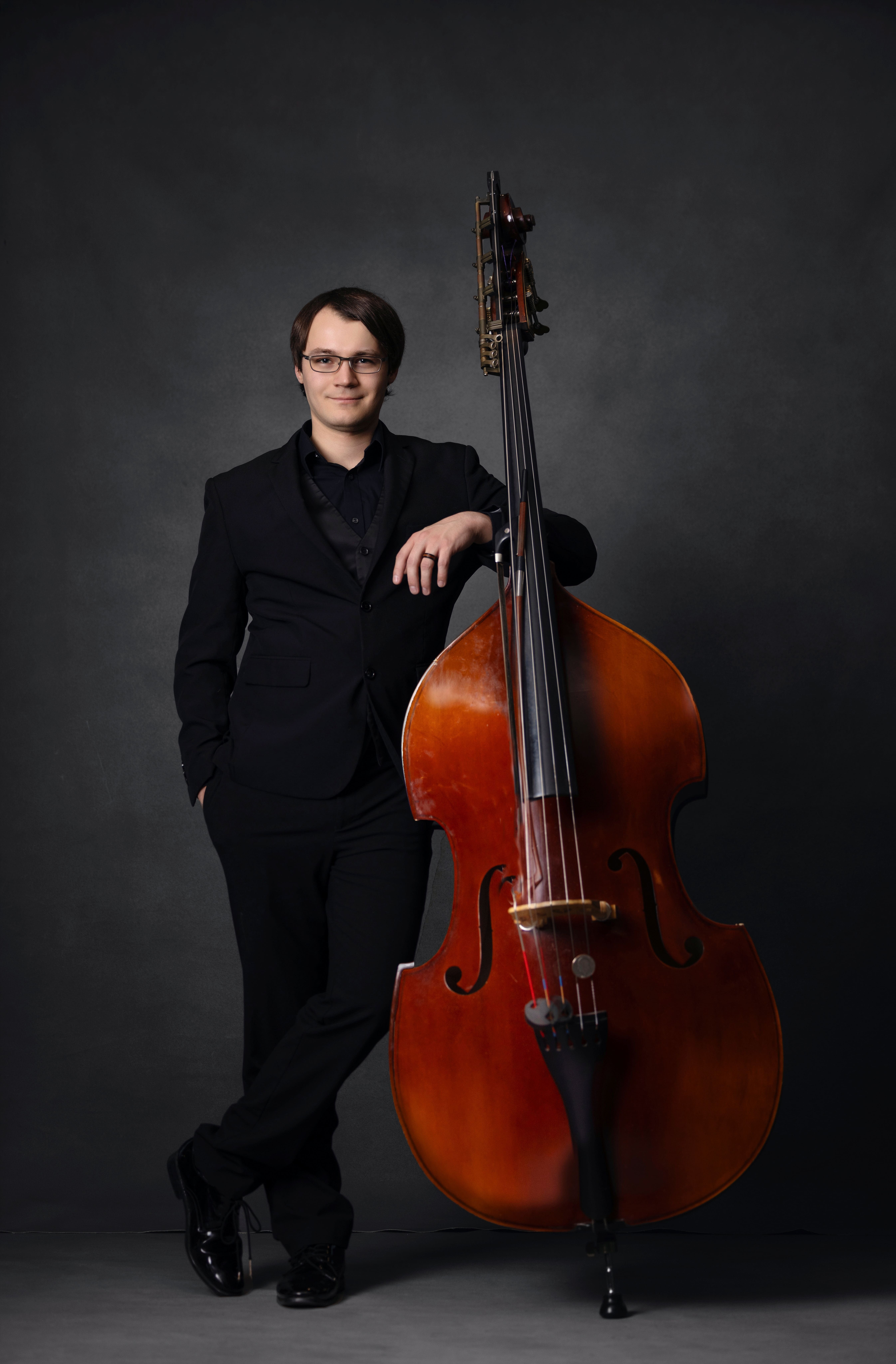 Male musician in a black suit leaning on a wooden double bass against a dark studio backdrop, wearing glasses.