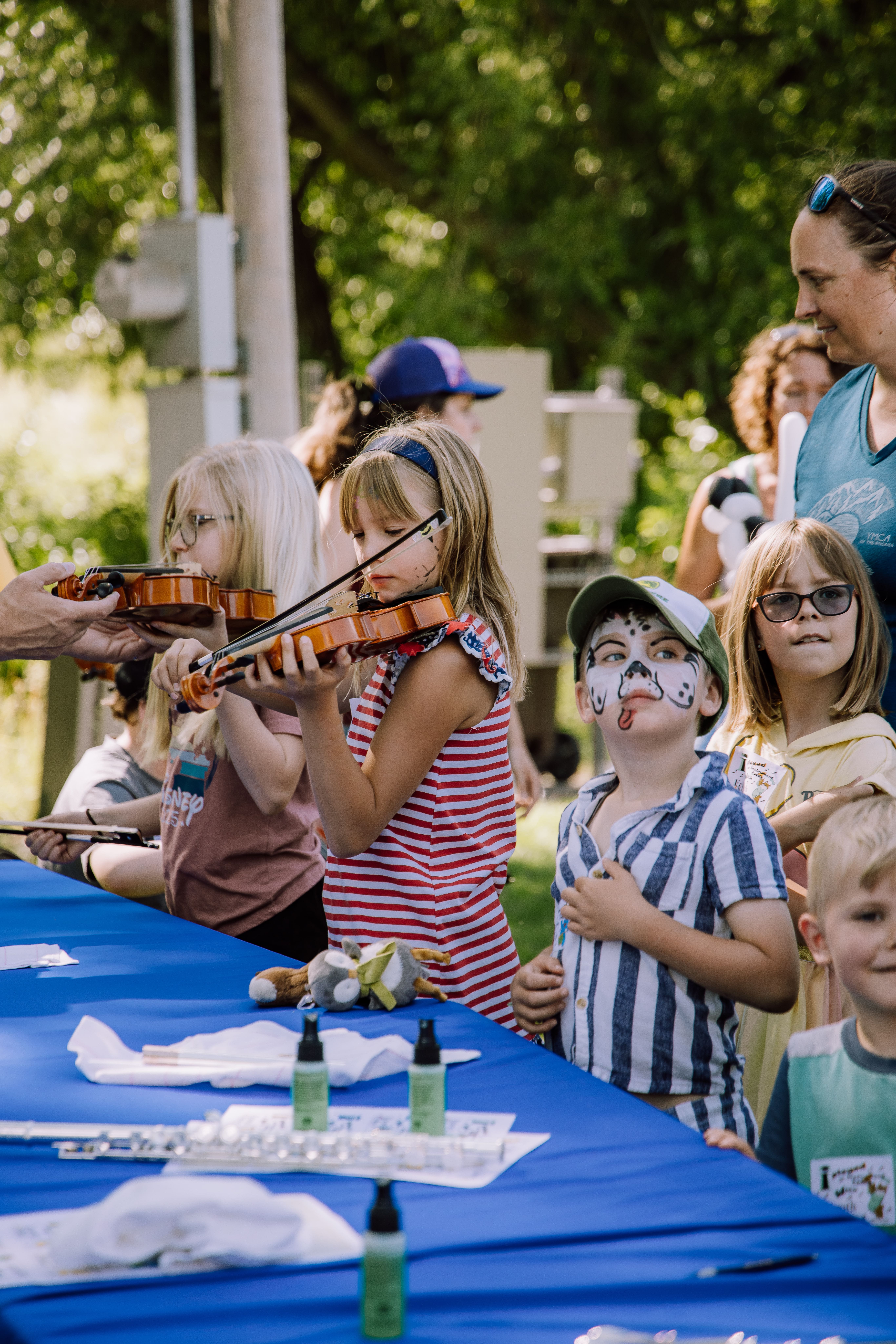 Group of children playing violins outdoors at a sunny event, standing by a blue table with craft supplies nearby.