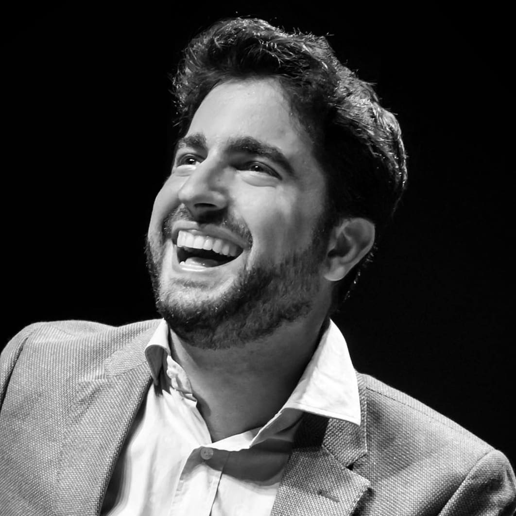 Portrait of a smiling man with dark hair and beard, wearing a light blazer and white shirt, laughing against a dark background