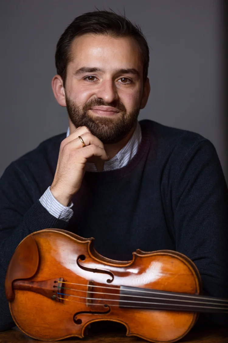 Portrait of a man with a beard in a navy sweater, resting his chin beside a violin.