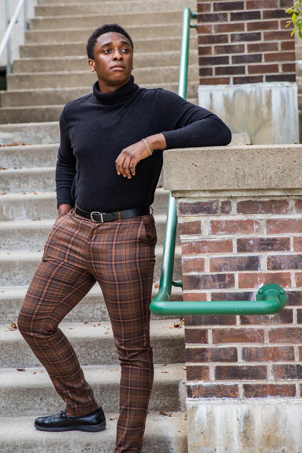Young man leaning on a brick railing beside concrete stairs, wearing a black turtleneck and brown plaid pants on a campus-style outdoor setting.