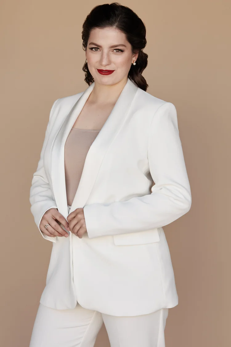 Woman in a white blazer and matching pants posing against a beige background, wearing neutral top and red lipstick.