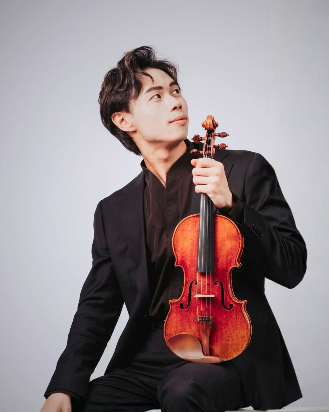 Young man in a black suit holding a violin, looking upward thoughtfully.