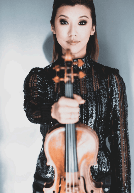 Female violinist in a sparkly black outfit extends a violin toward the camera, portrait shot with a soft background.