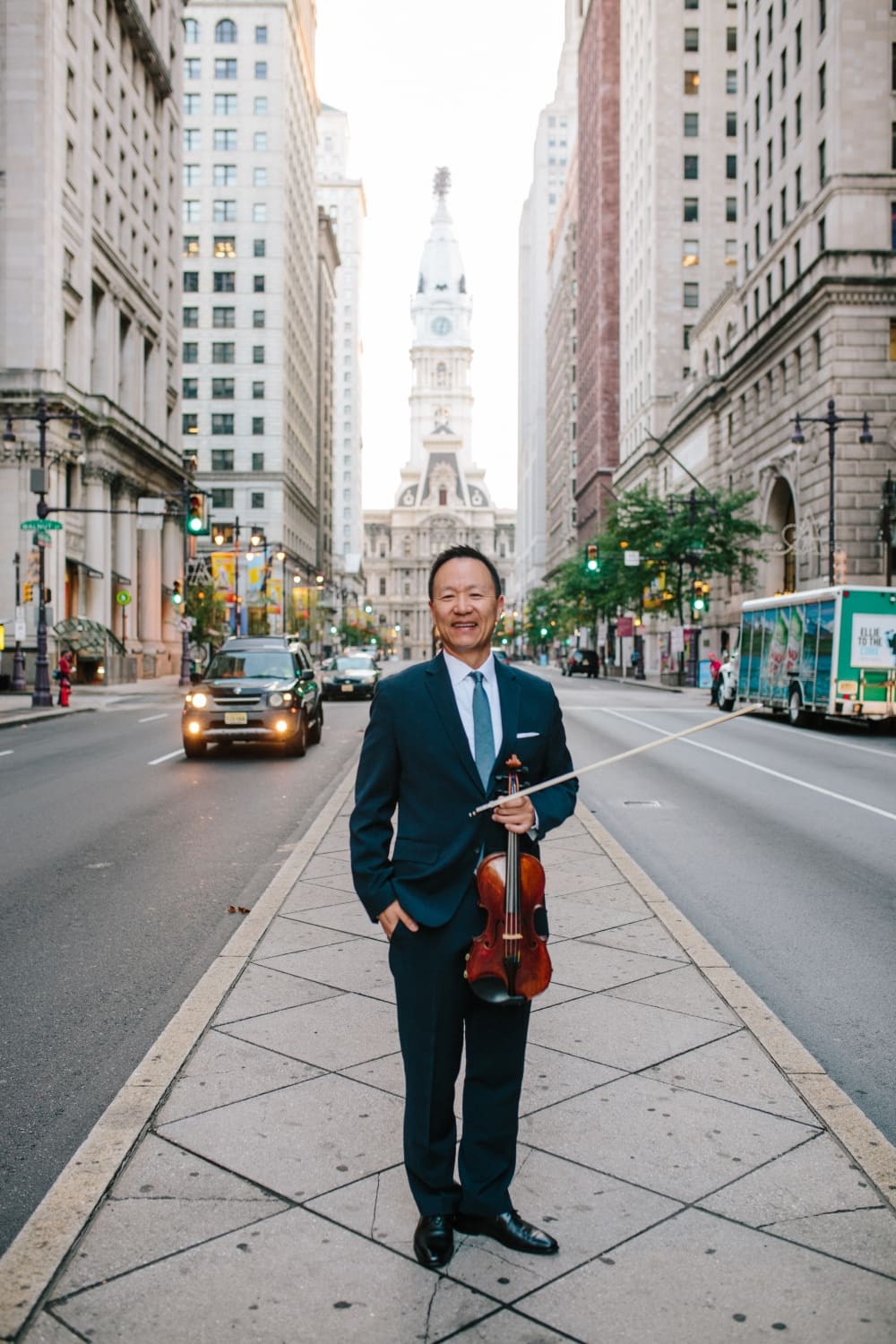 Smiling man in a navy suit holding a violin and bow on a city sidewalk with tall buildings and traffic behind him