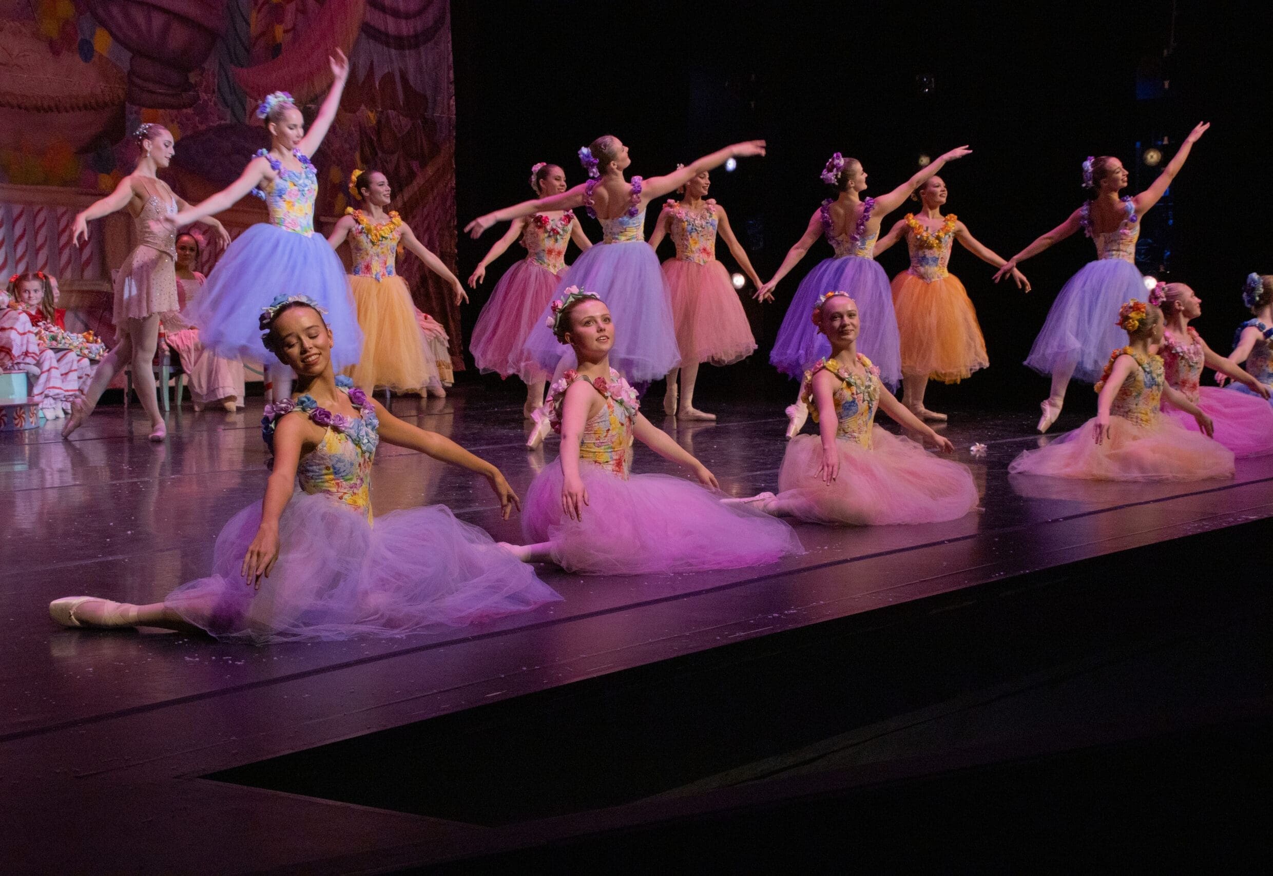 Group of young ballet dancers in colorful tutus performing on a stage with a decorative backdrop and floral headpieces.