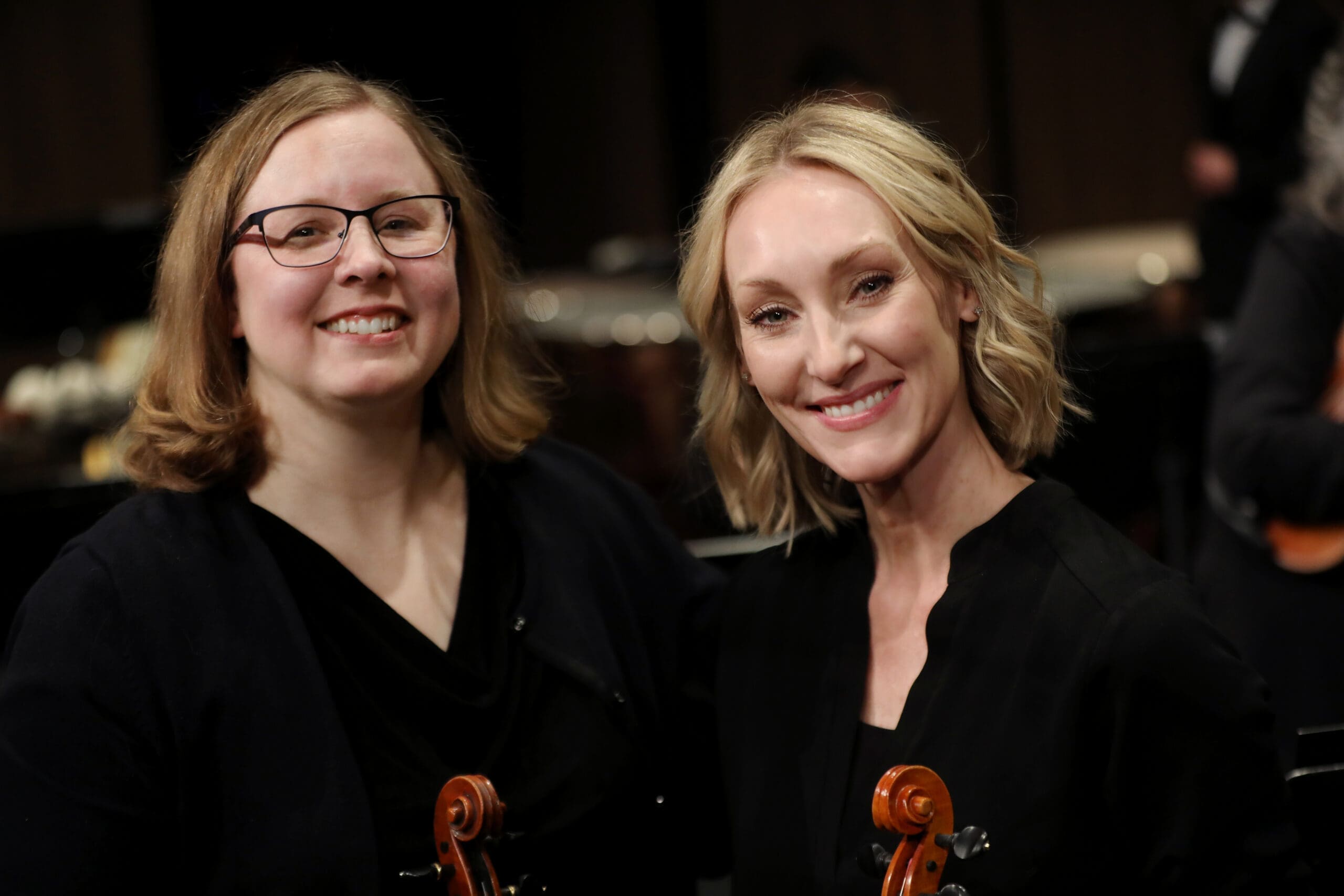 Two women in black, smiling at the camera, each holding a violin on a stage or backstage setting.