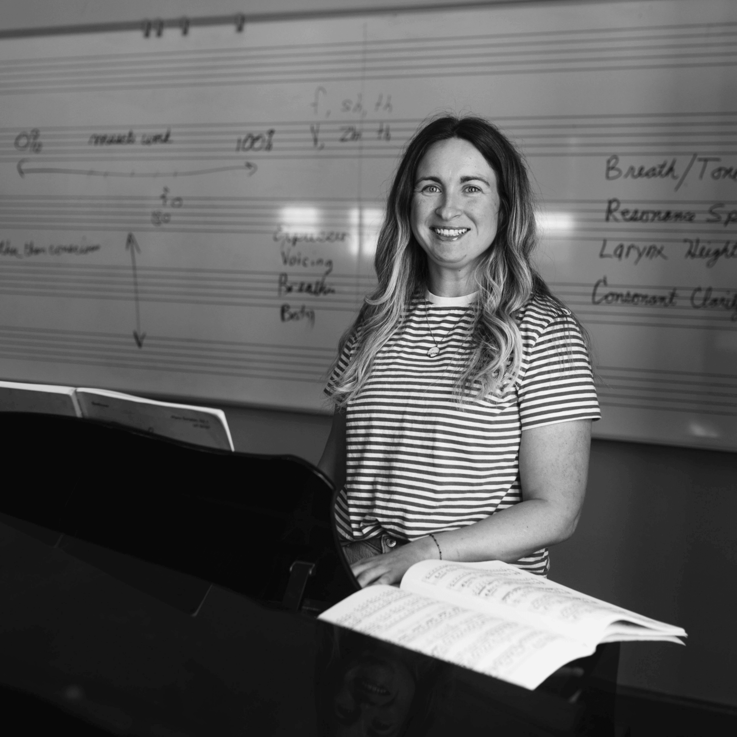 Smiling woman sits at a grand piano with open sheet music, a staff-covered whiteboard in the background, black-and-white photo.